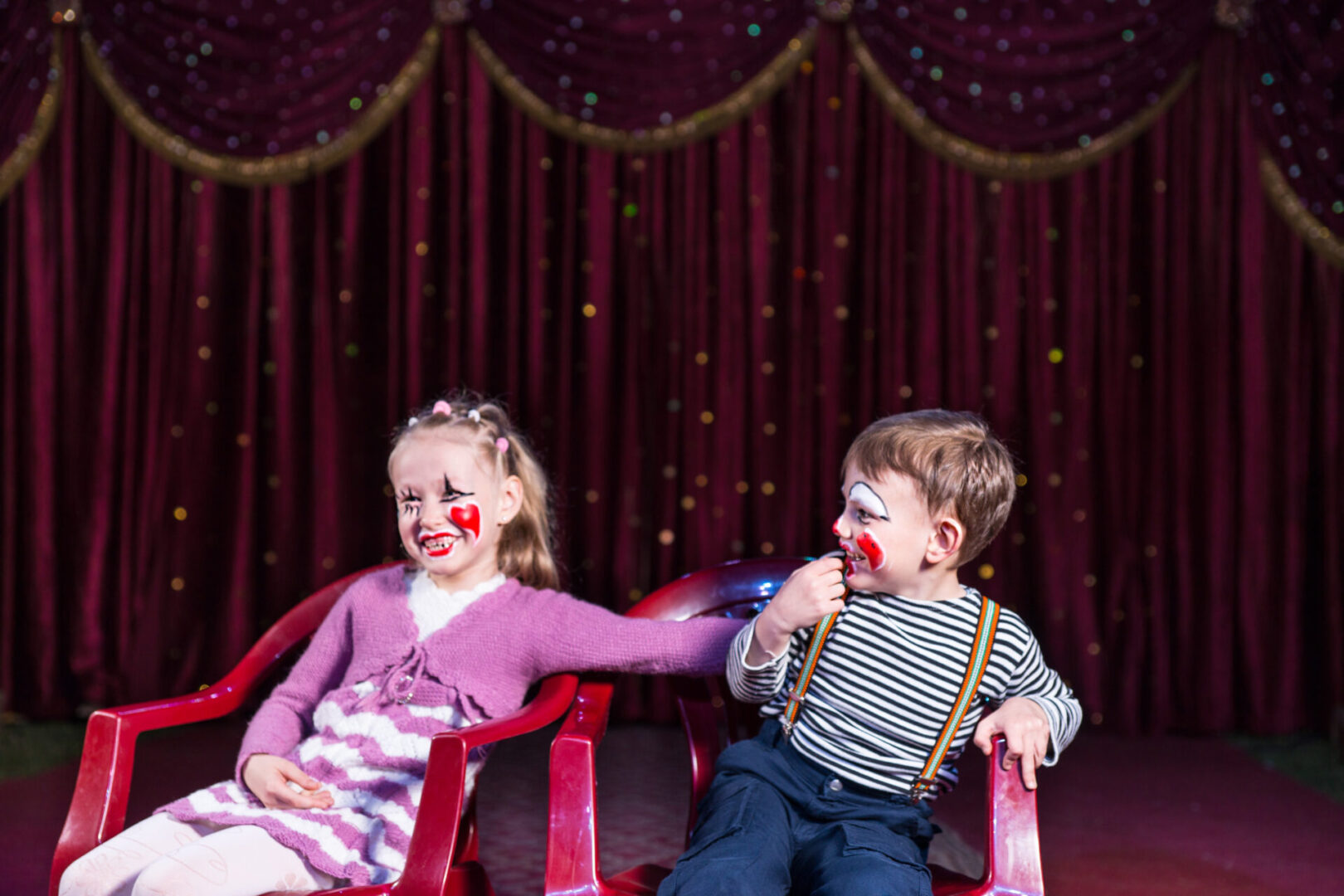 Girl and Boy Wearing Clown Make Up Laughing and Playing Together Sitting on Red Chairs on Empty Stage with Curtain in Background